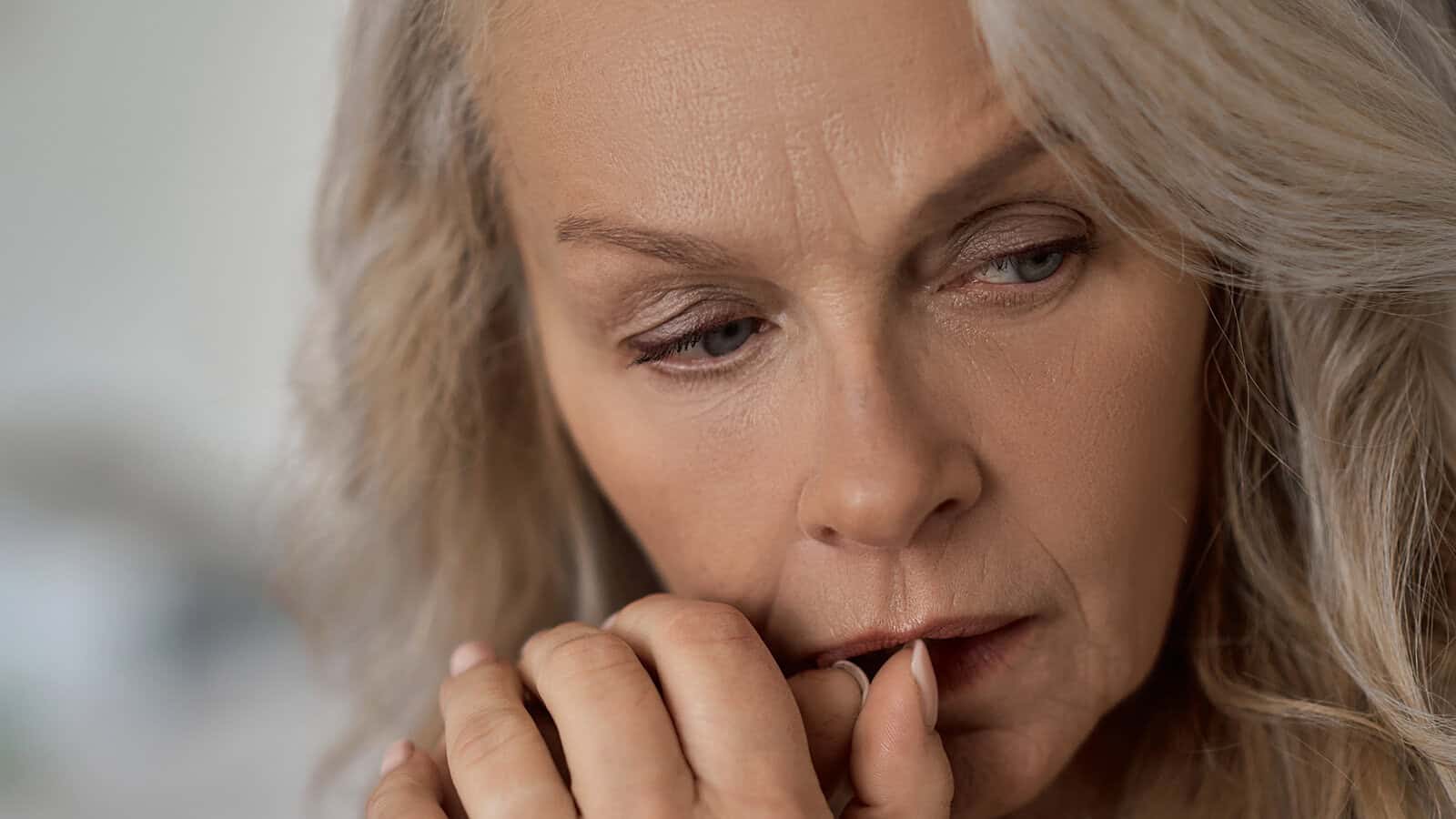 Close-up of a middle-aged woman with long gray hair, looking down with a worried expression and biting her nails, suggesting she is anxious or deep in thought.