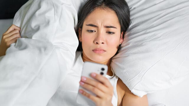 A woman lying in bed under white blankets looks at her phone with a concerned or confused expression.