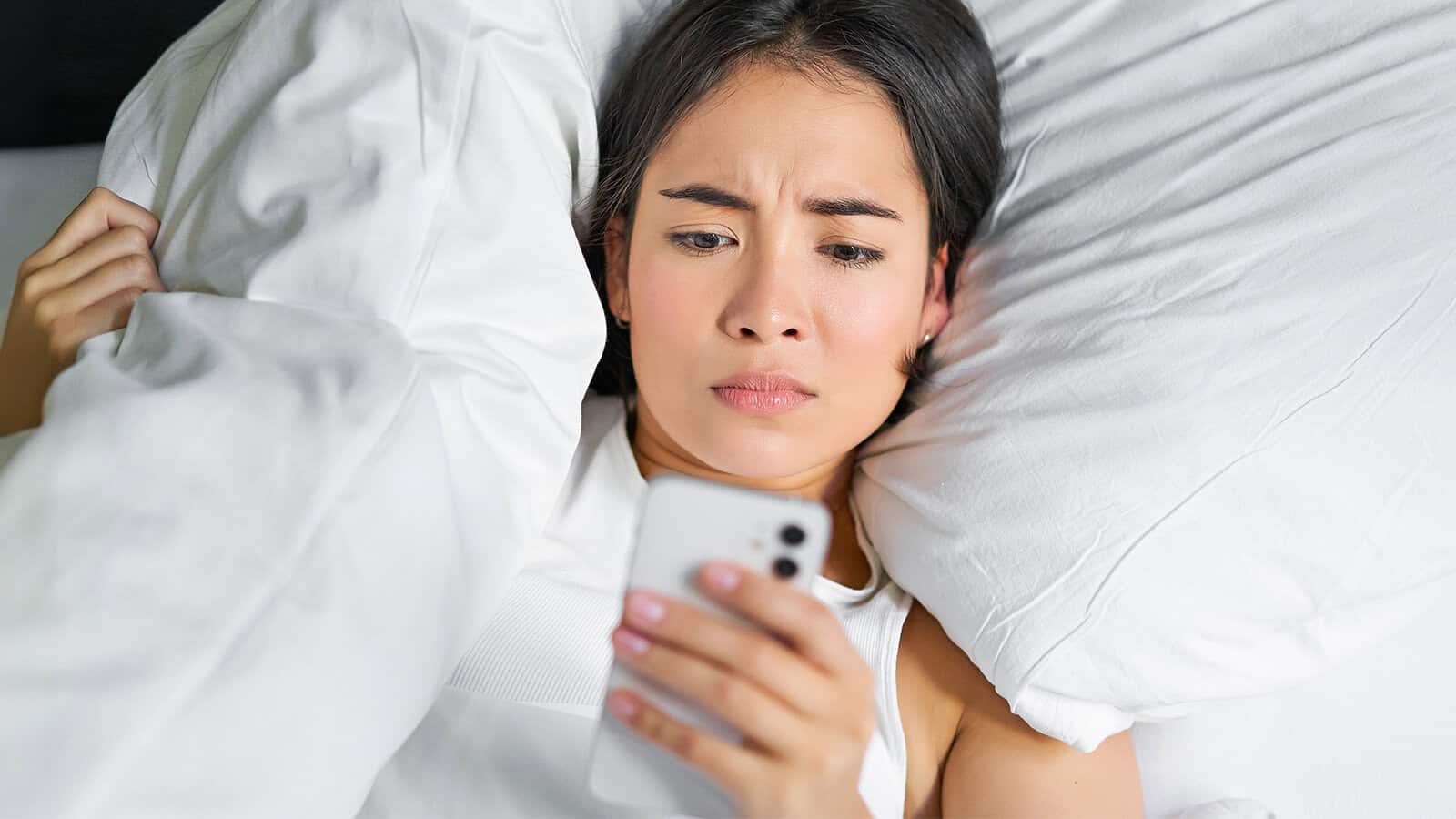 A woman lying in bed under white blankets looks at her phone with a concerned or confused expression.