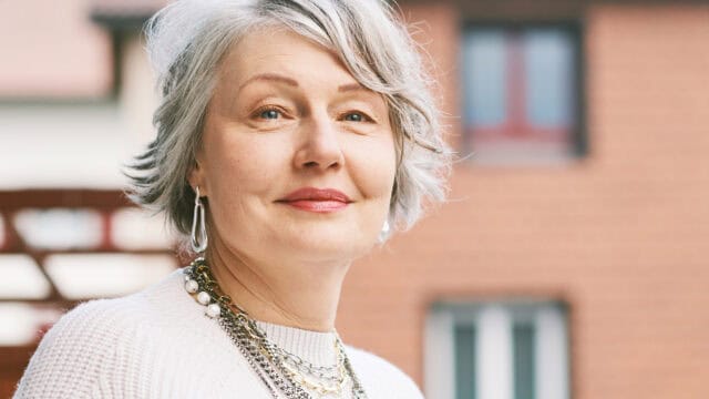 Smiling older woman with short gray hair and layered necklaces, wearing a white sweater, stands outdoors in front of blurred brick and cream-colored buildings.