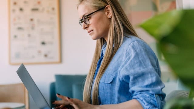 A woman with long blonde hair and glasses types on a laptop while sitting indoors. She wears a blue shirt and appears focused. The background includes a blurred poster and green plant.