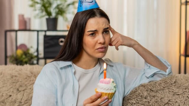A woman wearing a birthday hat looks sad while holding a cupcake with a lit candle, sitting on a couch in a cozy living room, quietly reflecting on her emotional needs during her birthday.