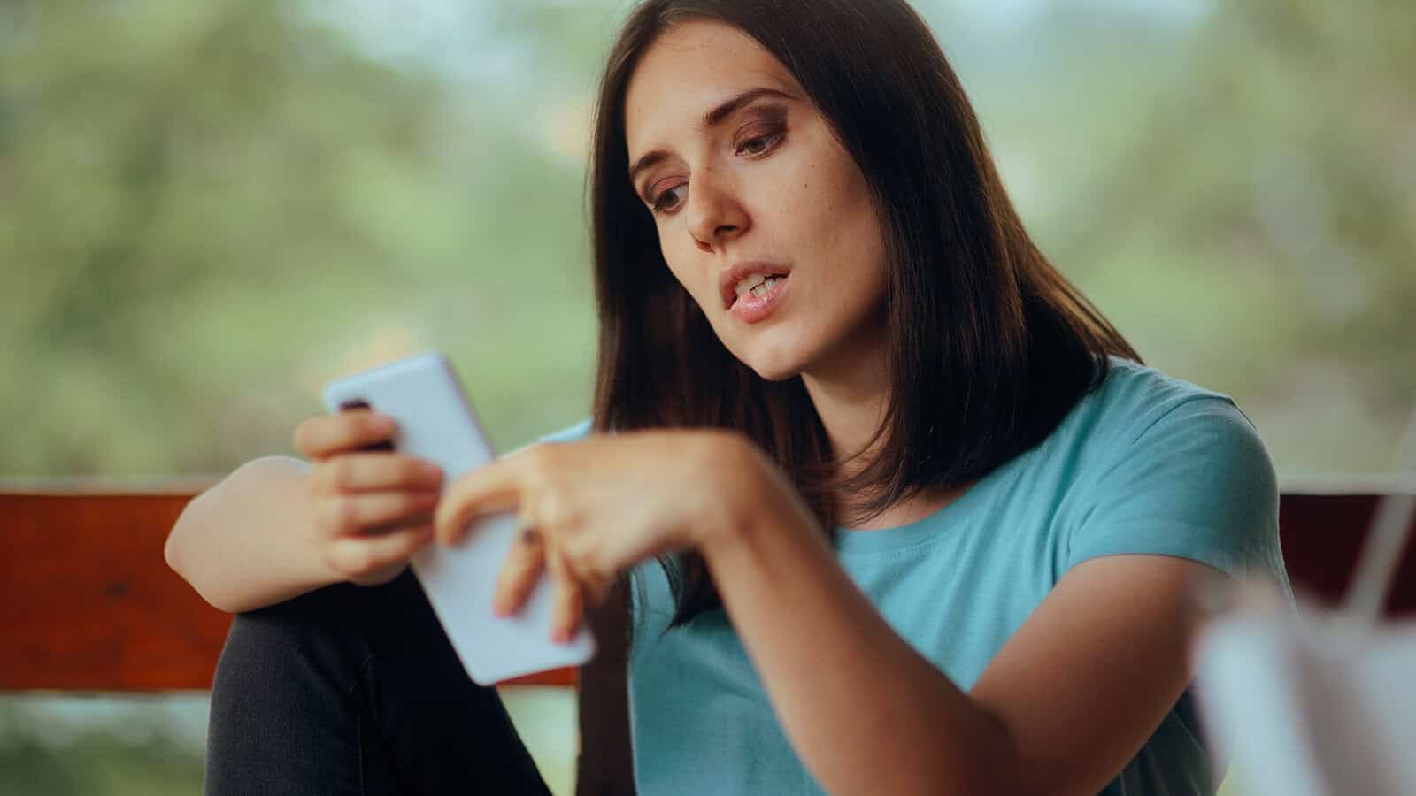 Young woman with long brown hair wearing a light blue shirt looks thoughtfully at her smartphone while sitting indoors, with a blurred green outdoor background.