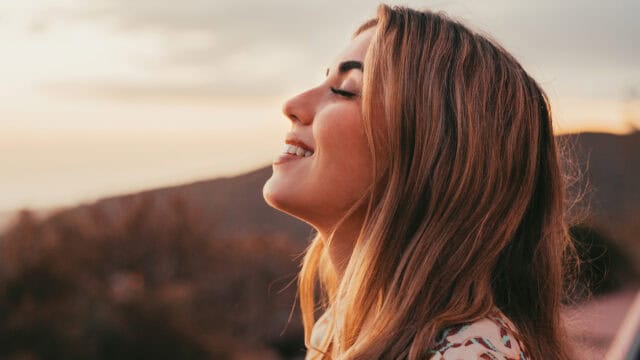 A woman with long hair smiles with her eyes closed, enjoying the outdoors at sunset. The background is blurred, showing warm sunlight and natural scenery.