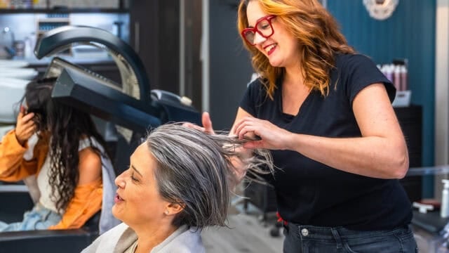 A hairstylist with red hair and glasses works on a smiling womans short gray hair in a modern salon. Another person sits under a hair dryer in the background.