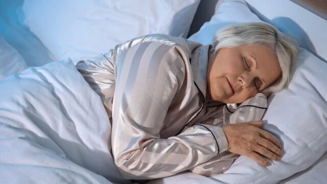An older woman with short gray hair sleeps peacefully on her side in bed, wearing light-striped pajamas and covered with a white blanket.