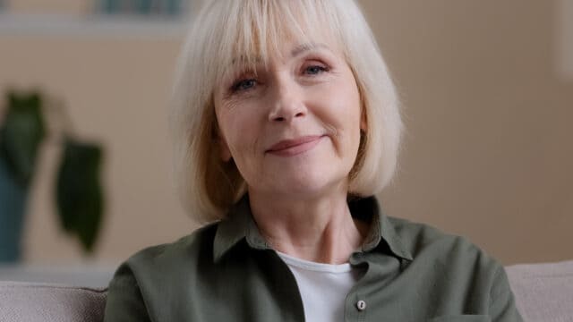 An older woman with short, light blonde hair and a gentle smile sits indoors on a couch, wearing a green shirt over a white top. A leafy plant is blurred in the background.