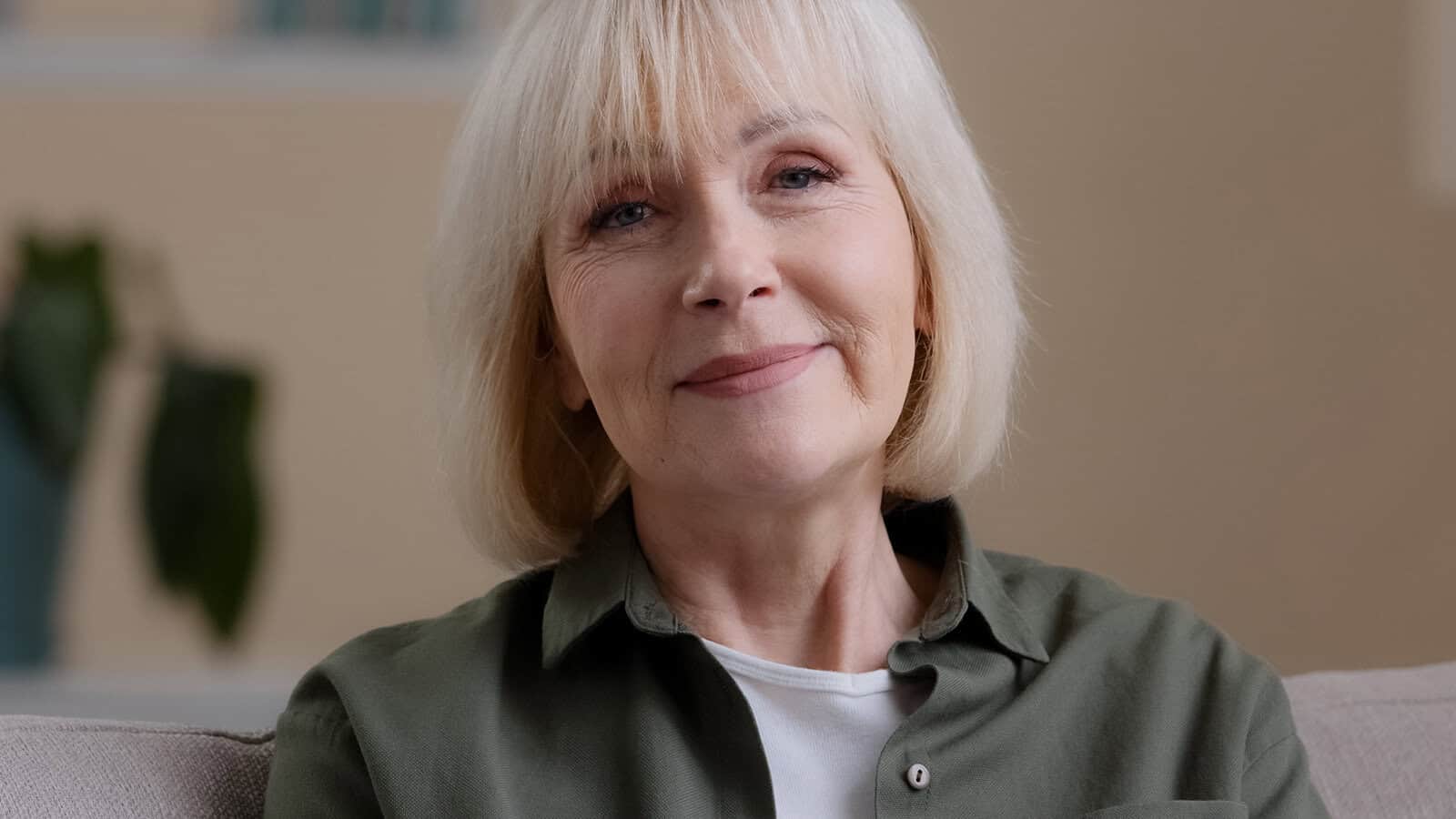 An older woman with short, light blonde hair and a gentle smile sits indoors on a couch, wearing a green shirt over a white top. A leafy plant is blurred in the background.