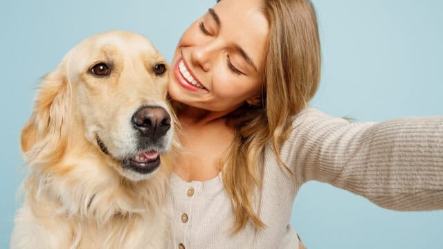 A smiling woman takes a selfie with a golden retriever against a light blue background. Both look happy and close together.