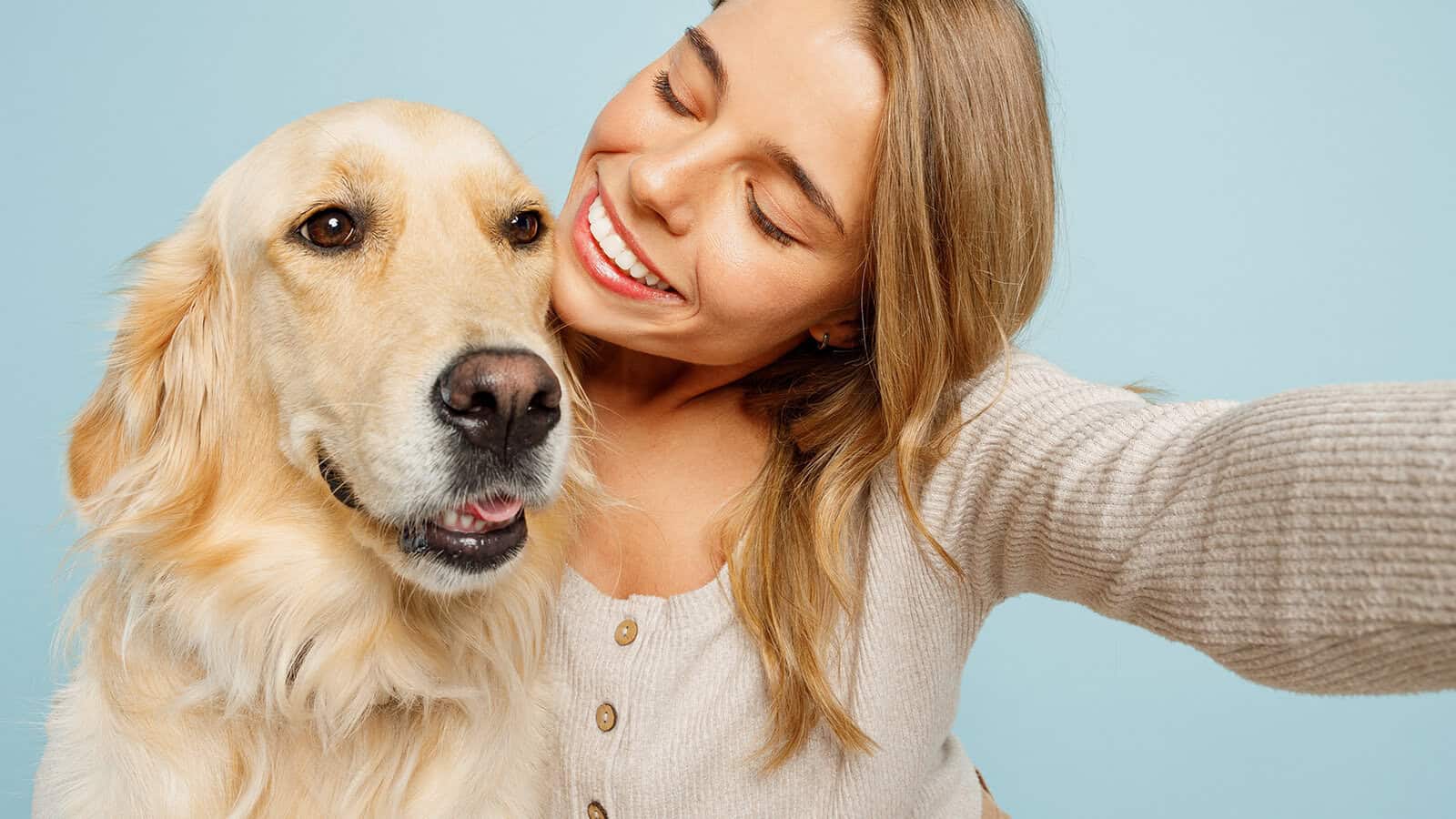 A smiling woman takes a selfie with a golden retriever against a light blue background. Both look happy and close together.