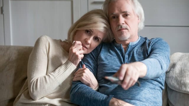An older couple sits on a couch, the woman leaning on the mans shoulder and holding his arm, looking emotional. The man holds a remote control and looks ahead, appearing calm and focused.