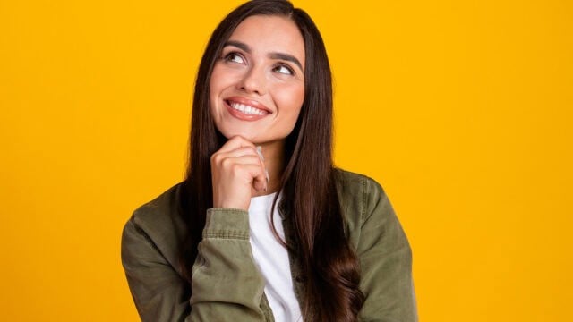 Woman with long brown hair, wearing a green jacket and white shirt, smiling and looking up thoughtfully against a bright yellow background.