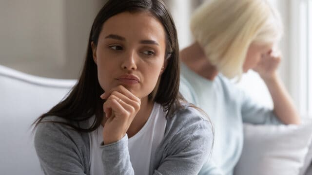 A young woman looks thoughtful and concerned while sitting on a couch, with an older woman in the background turned away, appearing upset.