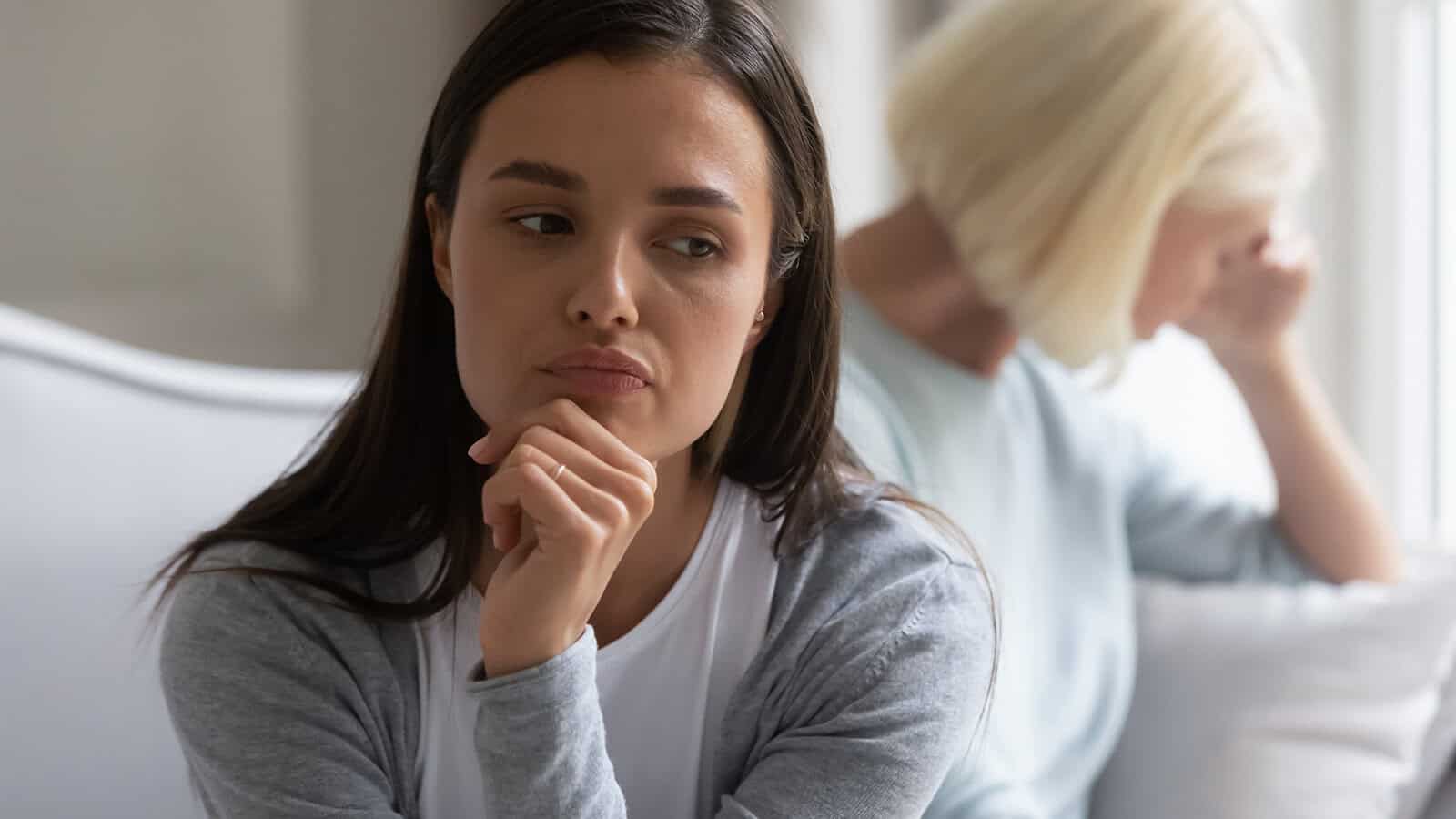 A young woman looks thoughtful and concerned while sitting on a couch, with an older woman in the background turned away, appearing upset.