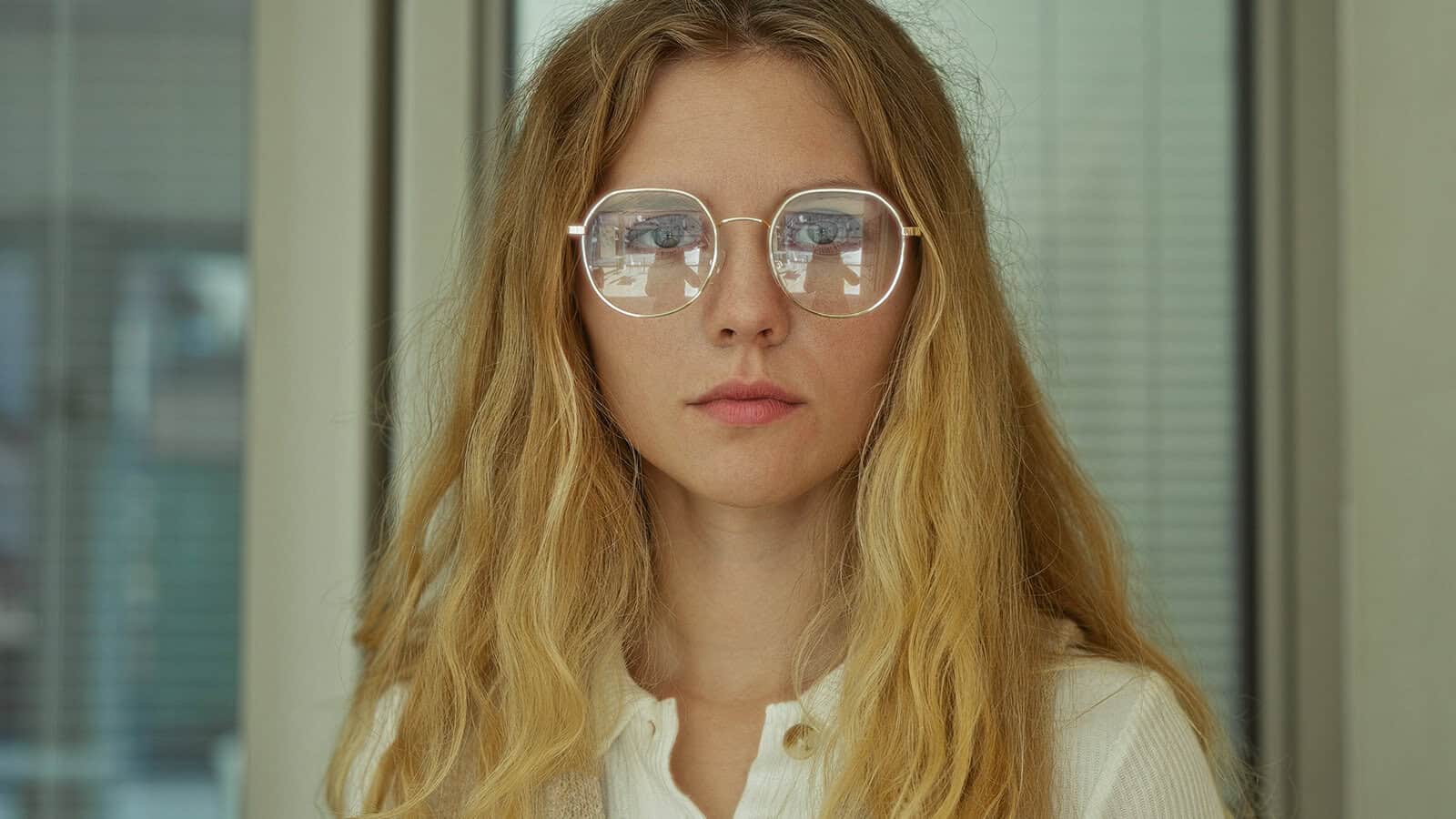 A young woman with long blonde hair and round glasses looks at the camera. She is wearing a white shirt, and the background shows windows and blinds in an indoor setting.