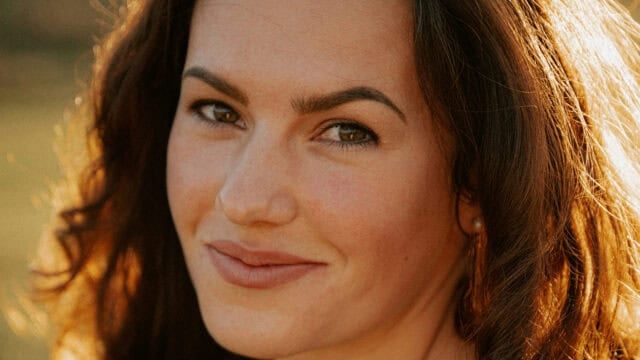 A woman with long brown hair smiles gently at the camera, sunlight highlighting her face and hair.