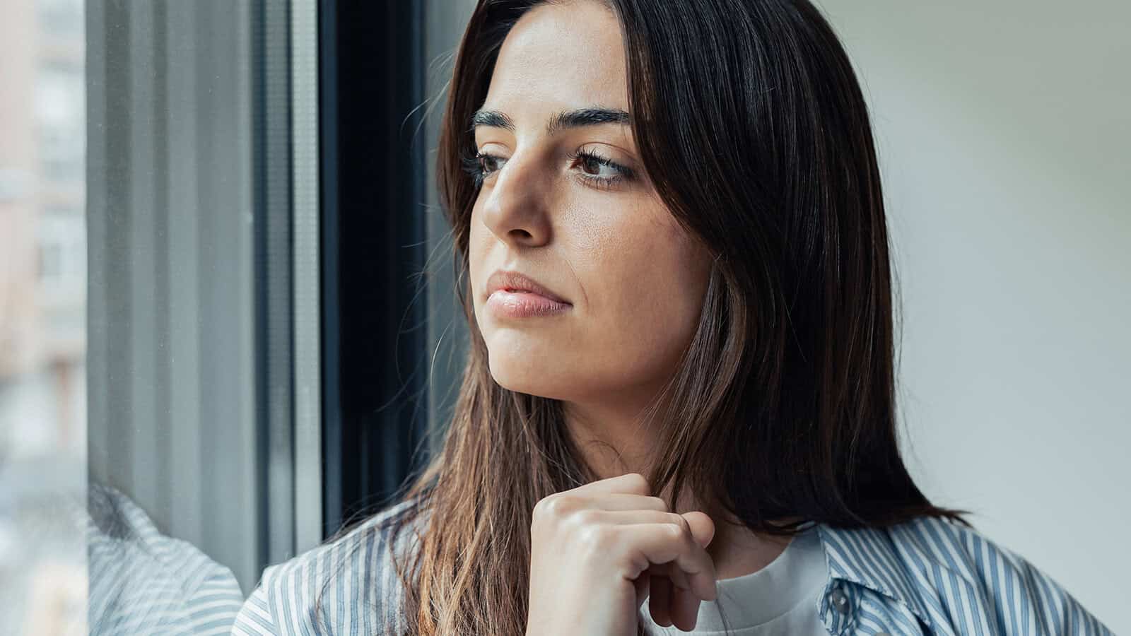 A woman with long brown hair looks thoughtfully out a window, resting her chin on her hand. She wears a striped shirt and appears contemplative, with natural light illuminating her face.