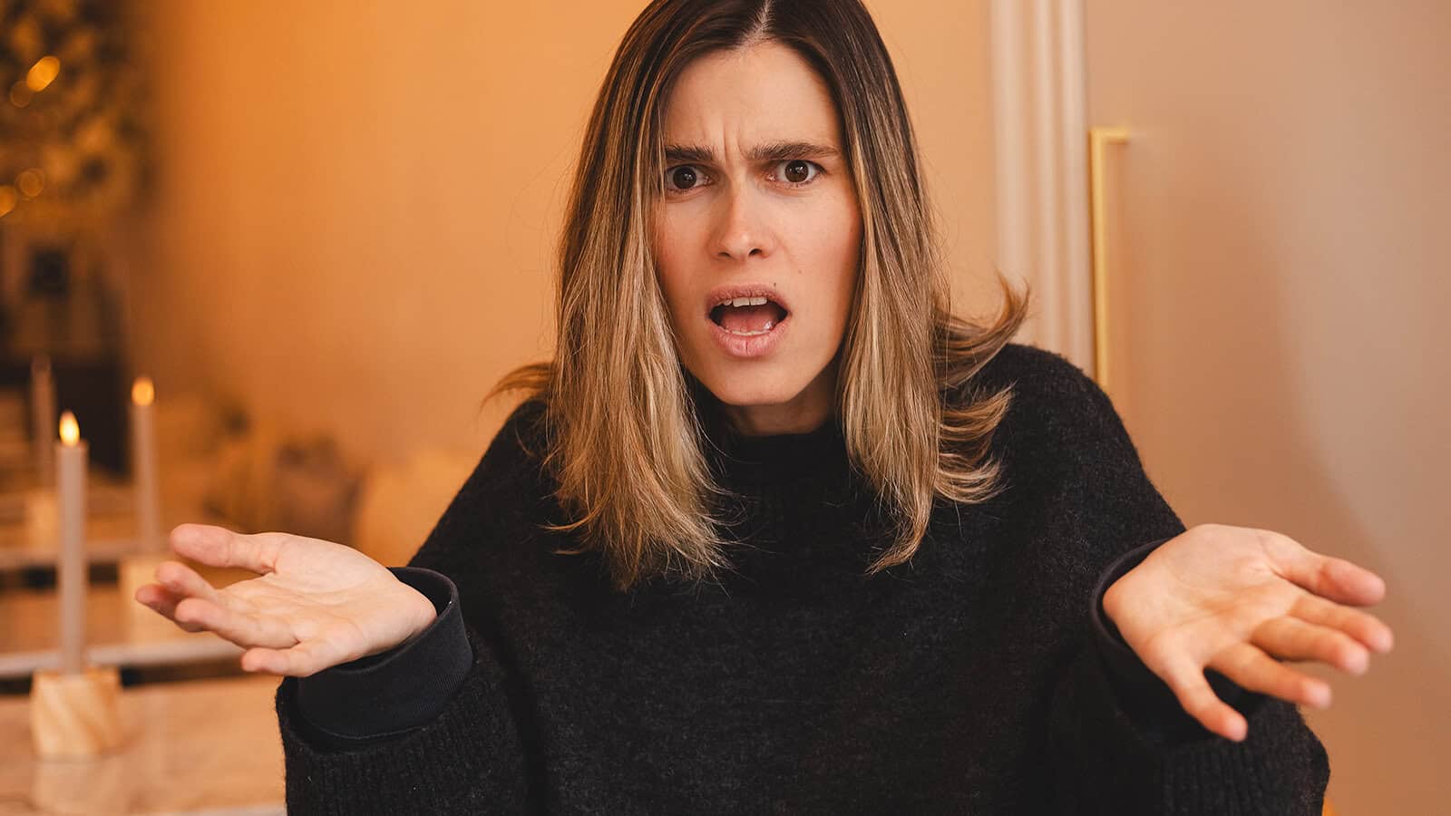A woman with shoulder-length hair sits indoors, looking surprised or confused, with her mouth open and both hands raised, palms up in a questioning gesture.