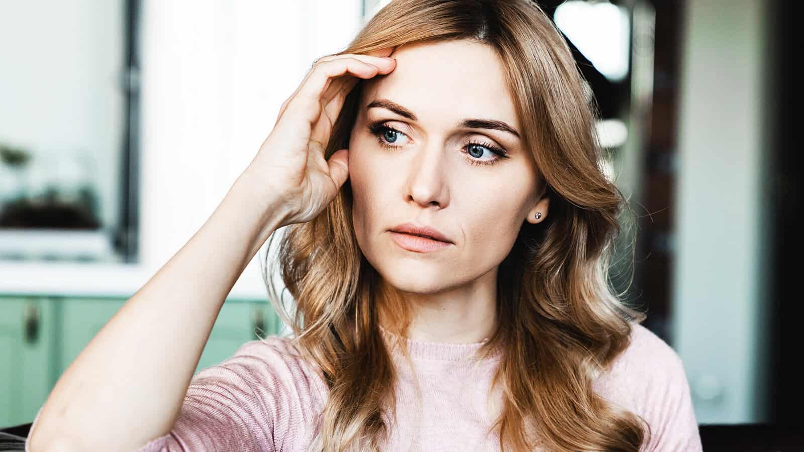 A woman with wavy blonde hair and a pink sweater looks thoughtful and concerned, resting her hand on her forehead, sitting indoors with a blurred background.