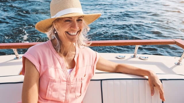 Smiling woman in a straw hat and pink shirt sits on a boat with water in the background, enjoying a sunny day.
