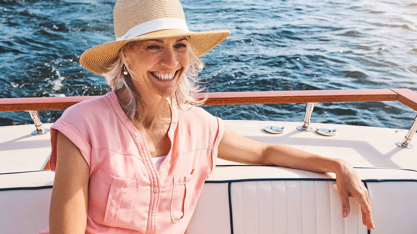 Smiling woman in a straw hat and pink shirt sits on a boat with water in the background, enjoying a sunny day.