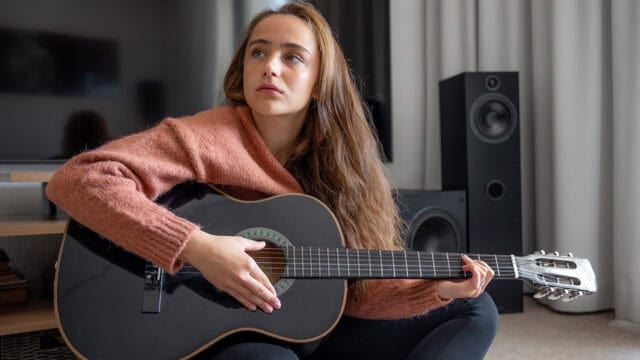 A young woman with long brown hair sits indoors, holding a black acoustic guitar and looking off to the side. She wears a pink sweater and is seated near speakers and a television.