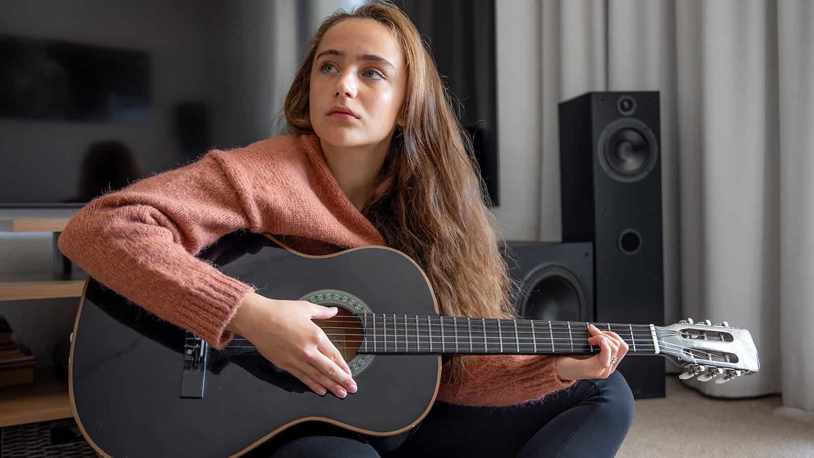 A young woman with long brown hair sits indoors, holding a black acoustic guitar and looking off to the side. She wears a pink sweater and is seated near speakers and a television.