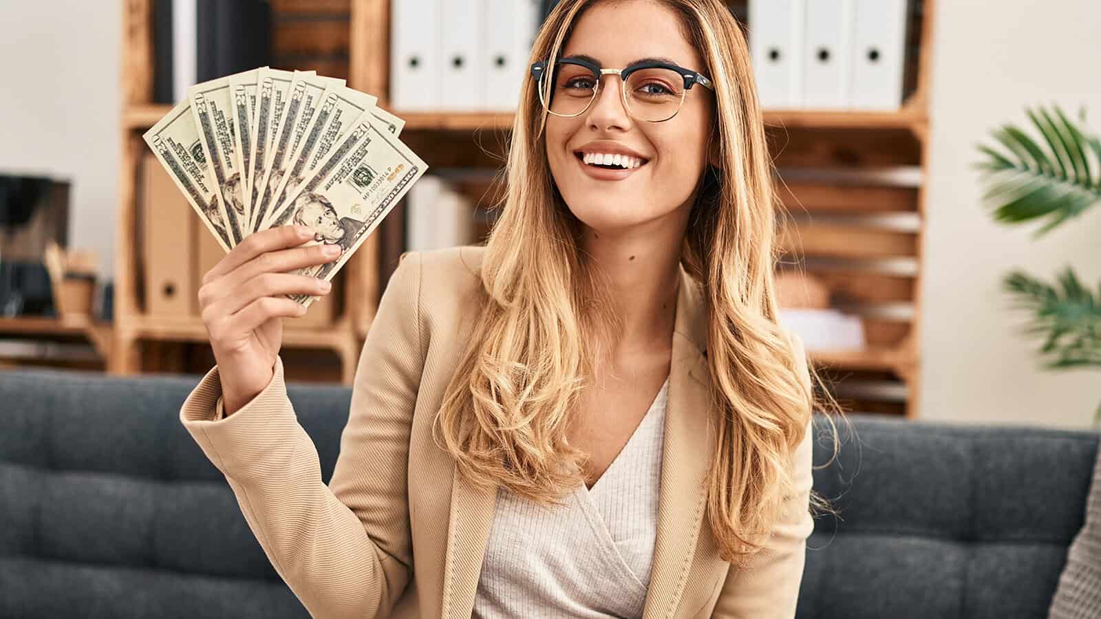 Smiling woman with long blonde hair and glasses sits on a sofa, holding a fan of US dollar bills in her hand. Office shelves and plants are visible in the background.