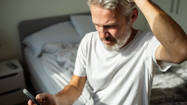 A man with gray hair and a beard sits on a bed in a bright room, looking at his smartphone with a confused or concerned expression while scratching his head.