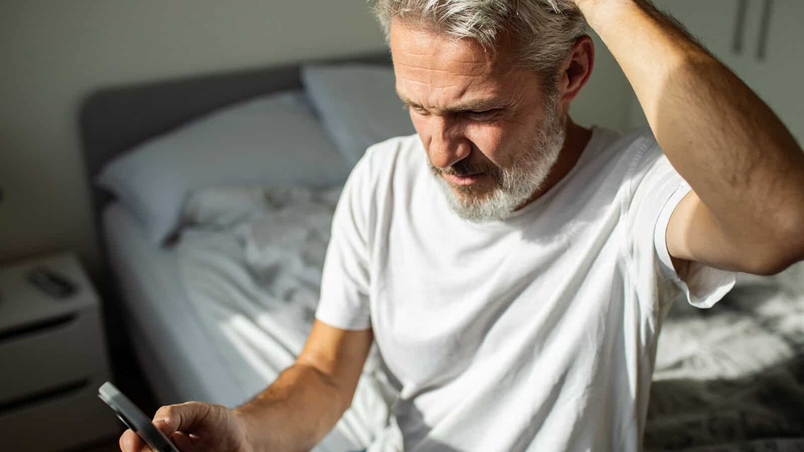 A man with gray hair and a beard sits on a bed in a bright room, looking at his smartphone with a confused or concerned expression while scratching his head.