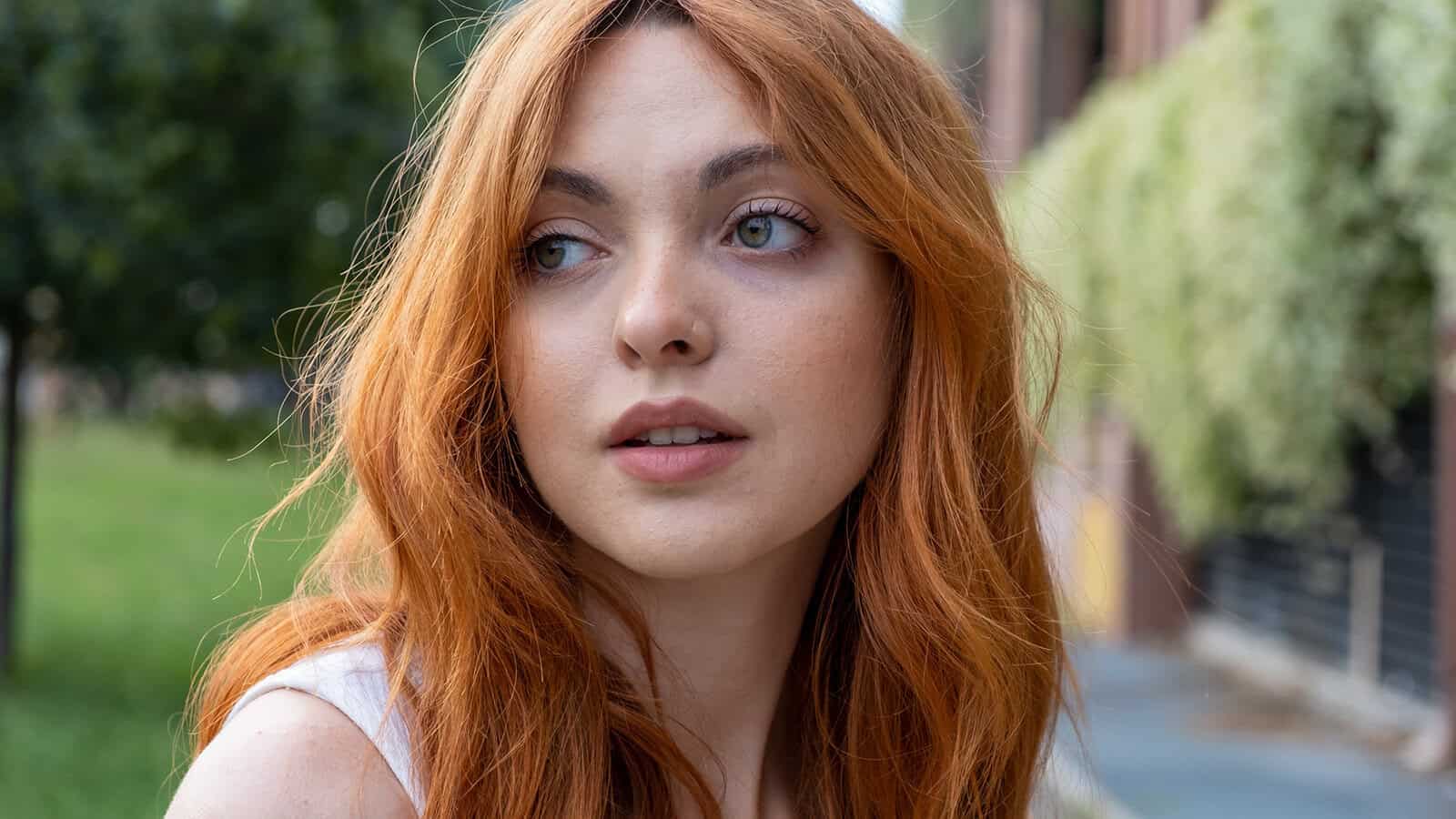 A woman with long red hair looks to her left while standing outdoors near greenery and buildings, with a thoughtful expression on her face.