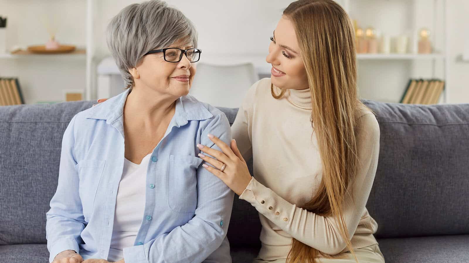 A young woman sits beside an older woman with gray hair on a couch, smiling and gently touching her shoulder in a cozy, softly lit living room.