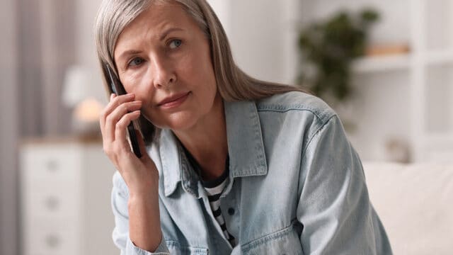 An older woman with gray hair, wearing a denim shirt, sits indoors and talks on a smartphone, looking thoughtful.