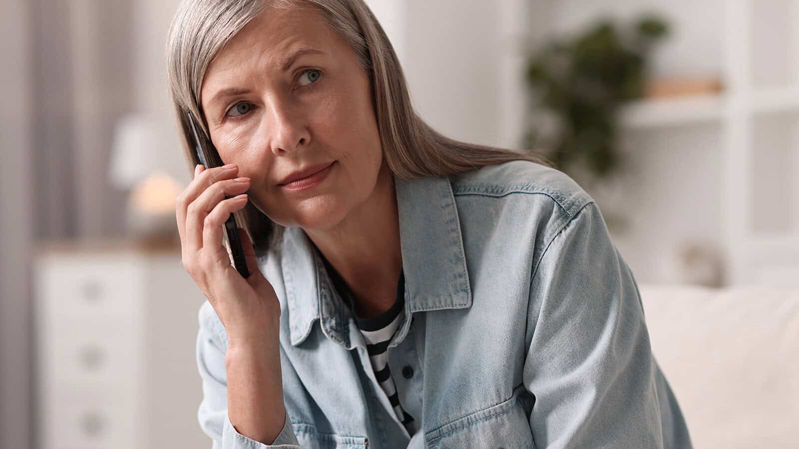 An older woman with gray hair, wearing a denim shirt, sits indoors and talks on a smartphone, looking thoughtful.