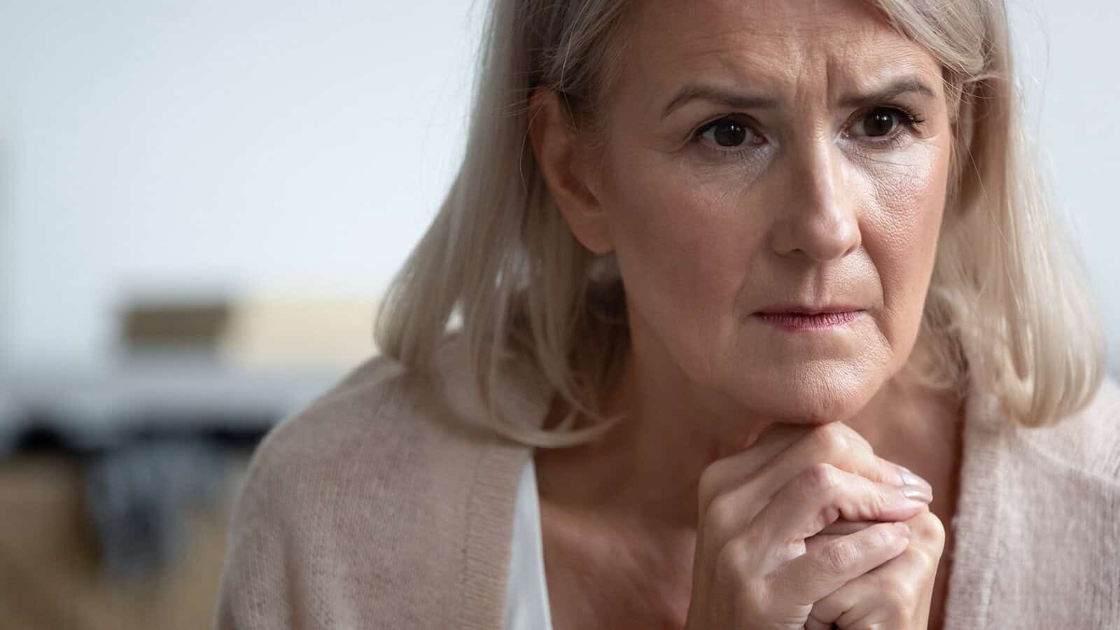 An older woman with light hair looks concerned and thoughtful, resting her chin on her hands while gazing ahead. She is indoors, wearing a light sweater.