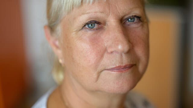 Close-up of an older woman with light skin, short blonde hair, and blue eyes, looking directly into the camera with a neutral expression. Indoor background is softly blurred.