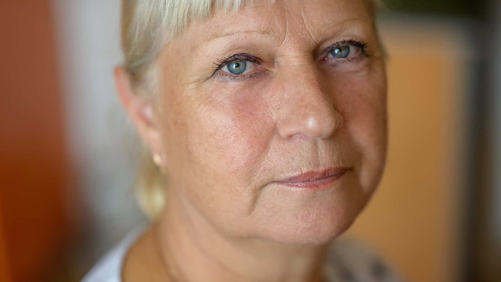 Close-up of an older woman with light skin, short blonde hair, and blue eyes, looking directly into the camera with a neutral expression. Indoor background is softly blurred.