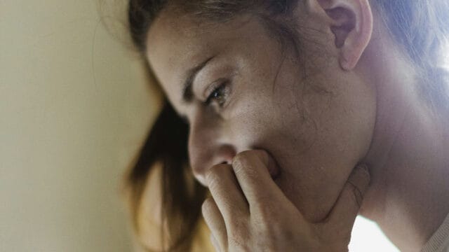 A person with brown hair appears deep in thought or worried, resting their chin on their hand and looking downward with a serious expression.