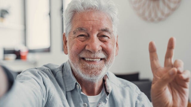 Smiling older man with gray hair and beard makes a peace sign with his hand while taking a selfie indoors.
