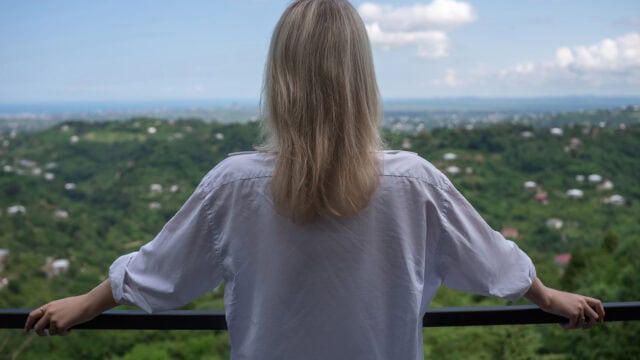 A person with long blonde hair, wearing a white shirt, stands on a balcony facing a green, hilly landscape dotted with houses under a blue sky with clouds.