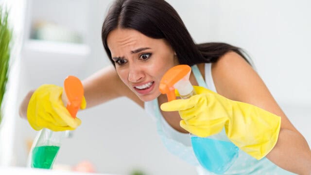 A woman wearing yellow cleaning gloves looks stressed while holding two spray bottles with different colored liquids, focusing intently as she cleans.