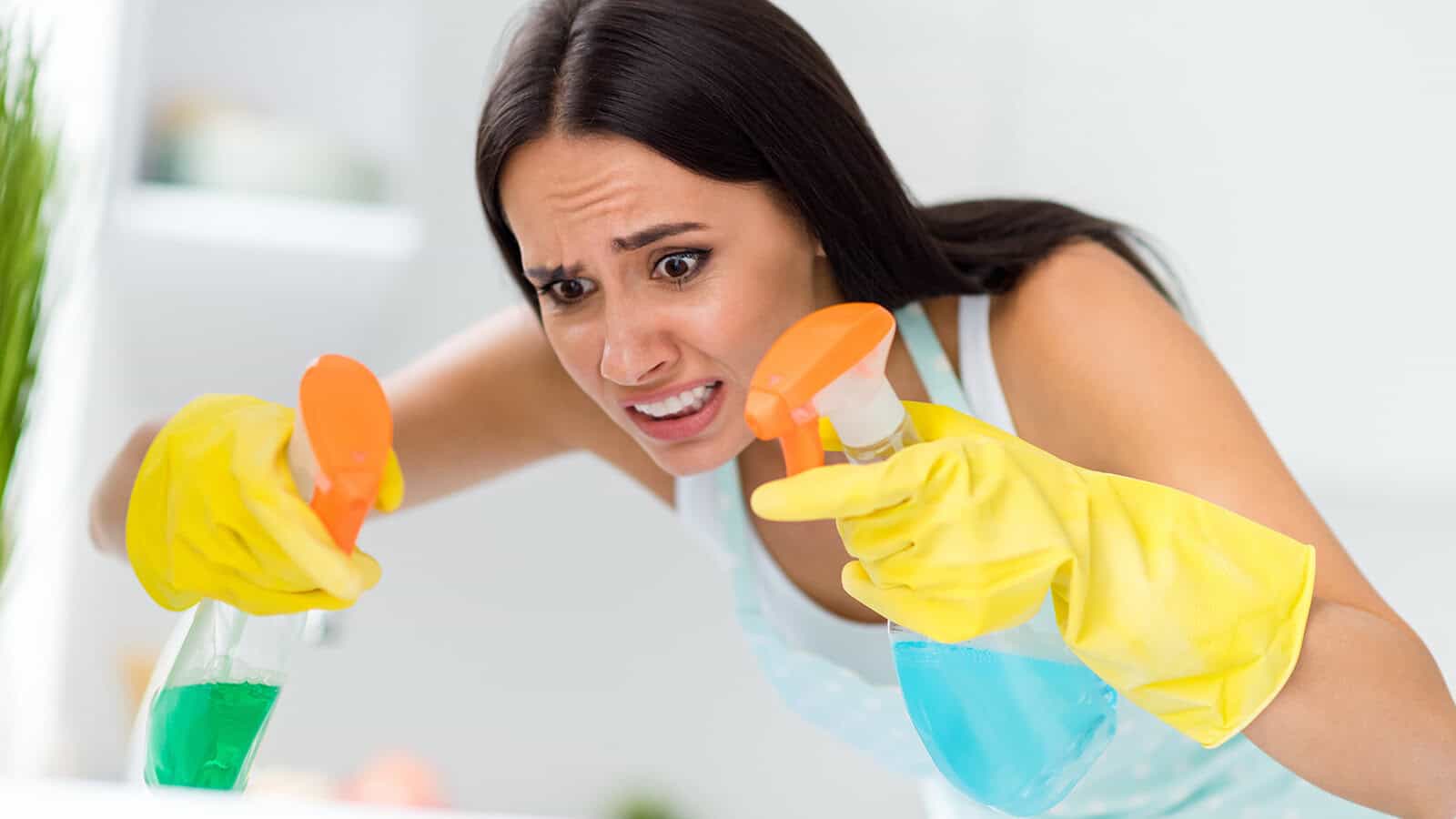 A woman wearing yellow cleaning gloves looks stressed while holding two spray bottles with different colored liquids, focusing intently as she cleans.