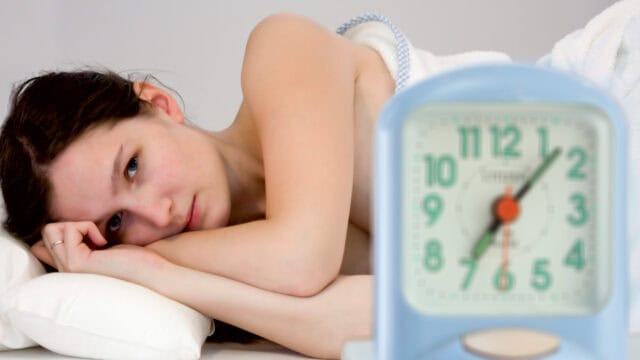A woman lies in bed with her head on a pillow, looking tired. A blurry alarm clock in the foreground shows just after 7:00.