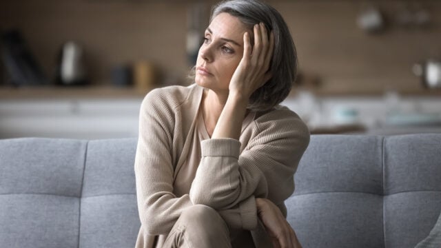 A woman with short gray hair sits on a couch, resting her head on her hand and looking off to the side, appearing thoughtful or concerned. She wears neutral-colored clothes in a softly lit room.