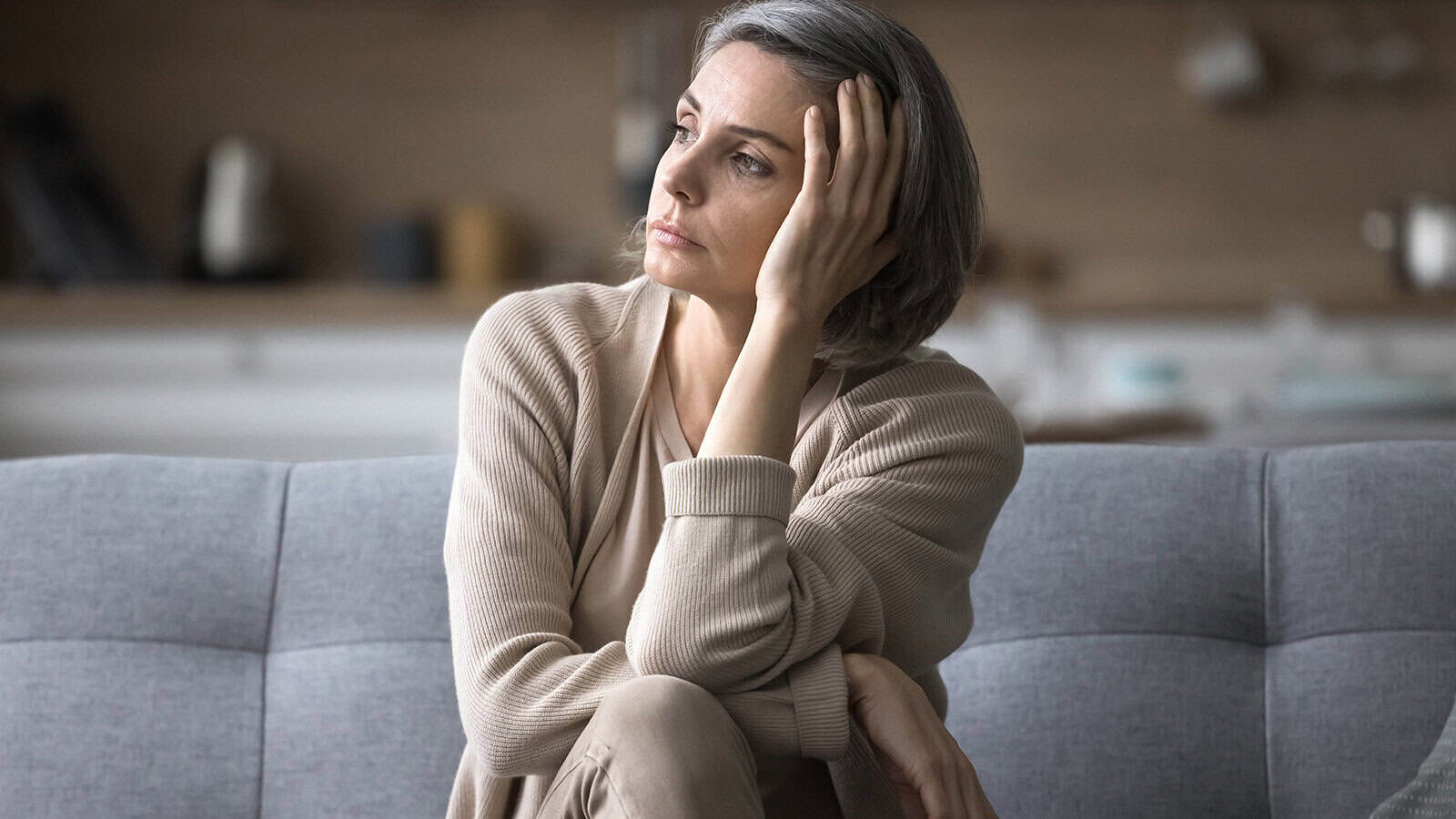 A woman with short gray hair sits on a couch, resting her head on her hand and looking off to the side, appearing thoughtful or concerned. She wears neutral-colored clothes in a softly lit room.