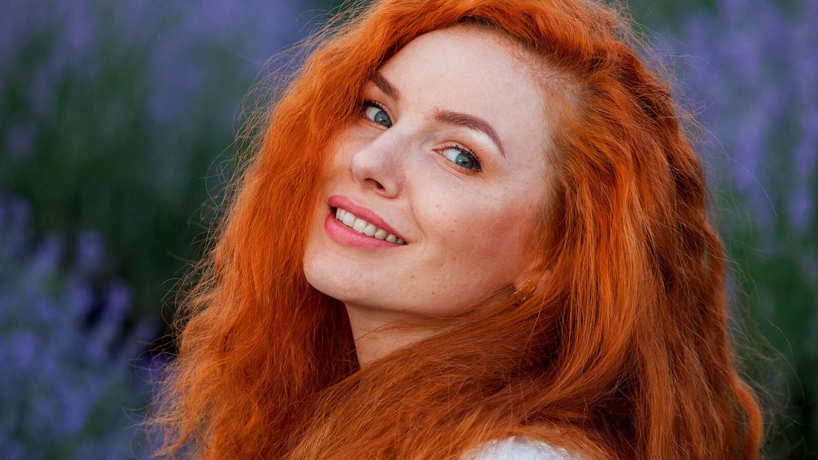 A person with long, curly red hair smiles while looking over their shoulder, standing outdoors with a blurred background of purple flowers or plants.