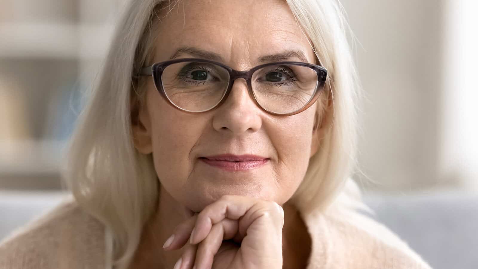 Older woman with long gray hair and glasses softly smiling, resting her chin on her hand, looking directly at the camera. She appears calm and thoughtful in a light, indoor setting.