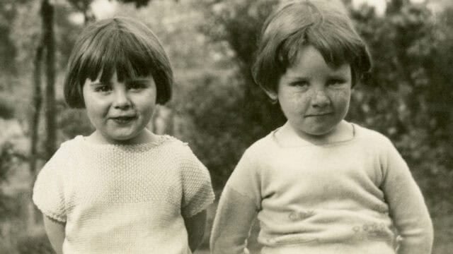 Two young children with short hair stand side by side outdoors, looking at the camera and smiling slightly. The image is in black and white, suggesting it is from an earlier era. Trees and foliage are visible in the background.
