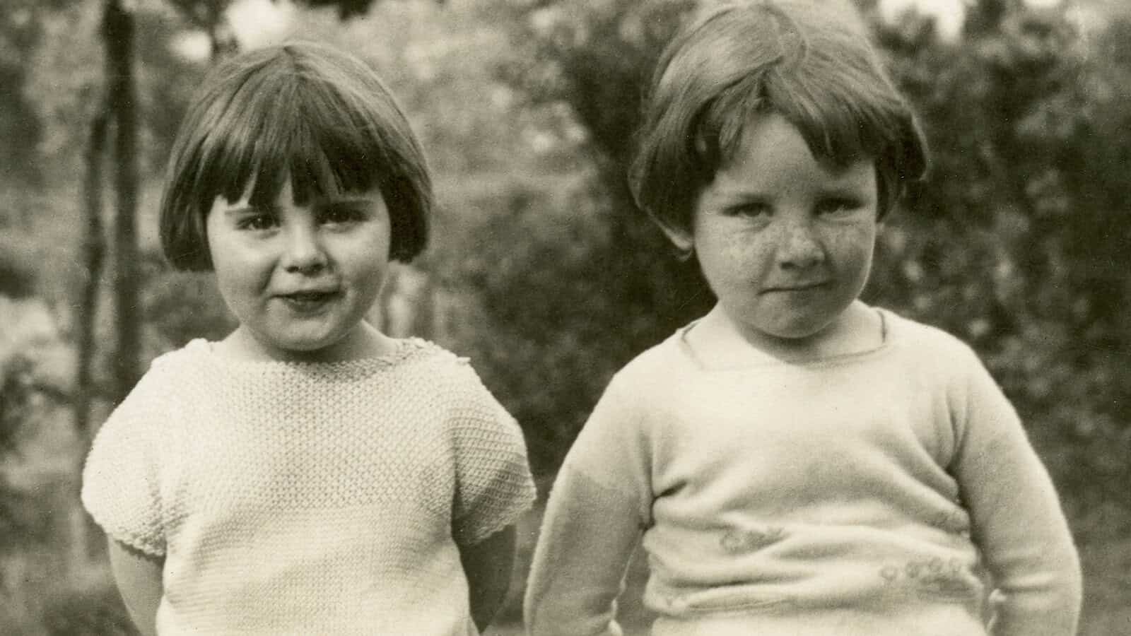 Two young children with short hair stand side by side outdoors, looking at the camera and smiling slightly. The image is in black and white, suggesting it is from an earlier era. Trees and foliage are visible in the background.