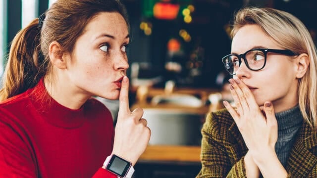 Two women sit together indoors; one in a red sweater holds her finger to her lips, signaling shh, while the other, wearing glasses and a brown jacket, looks at her with a surprised expression, covering her mouth with her hands.